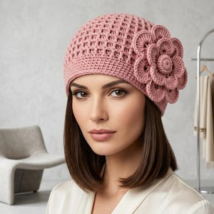 May include: A dusty rose crochet hat with a large, three-dimensional flower on the side. The hat has an open, lattice-like weave. The flower is made of layered, circular petals. The hat is a soft, feminine accessory.