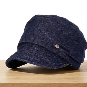May include: A dark blue denim cap with a short brim and a small, silver-colored button on the side. The cap is sitting on a wooden surface.