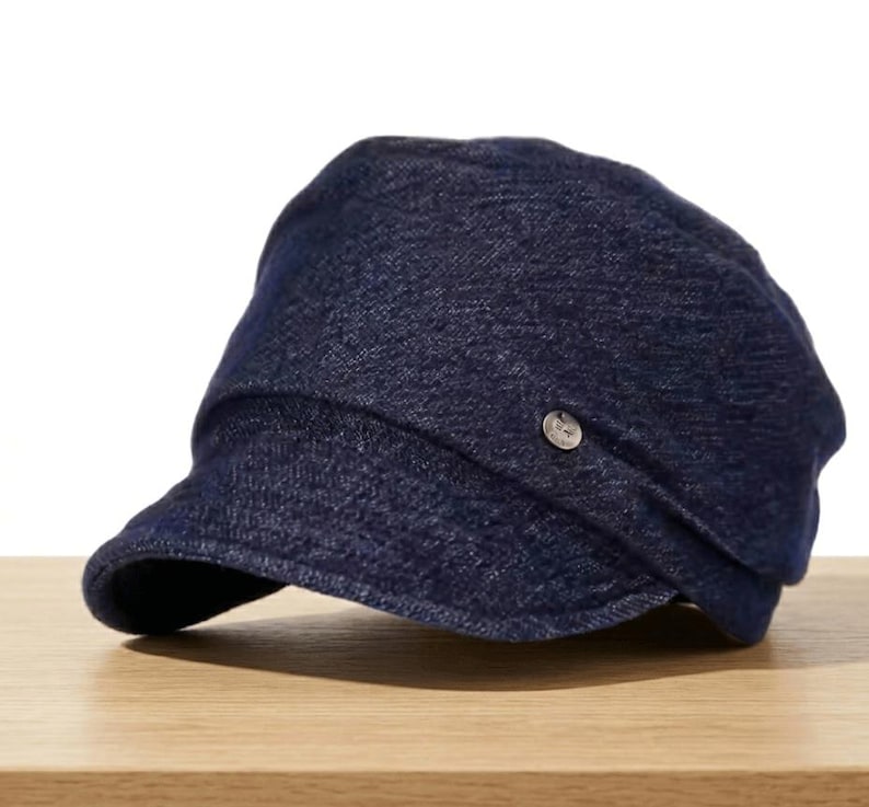 May include: A dark blue denim cap with a short brim and a small, silver-colored button on the side. The cap is sitting on a wooden surface.