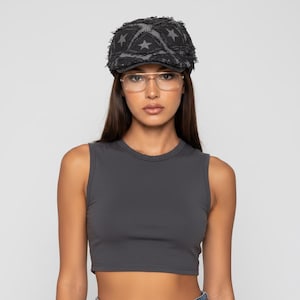 Op de afbeelding: Een donkergrijze pet met een versleten sterrenpatroon. De pet wordt gedragen met een grijze tanktop en een bril met gouden montuur. De algehele look is casual en stijlvol.