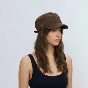 May include: Brown suede military-style cap with a short brim. The cap features decorative rivets on the side and a small strap detail at the back. The hat is a solid brown color and has a structured shape.