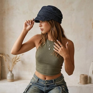 May include: A dark blue, textured newsboy cap with a curved brim. The cap is worn by a person wearing a green halter top and blue jeans. The person is also wearing a silver necklace and rings.