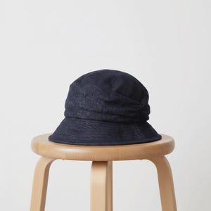 May include: A dark blue denim bucket hat with a rounded crown and a slightly wider brim. The hat is sitting on a light brown wooden stool against a white background. The hat has a gathered detail on the side.