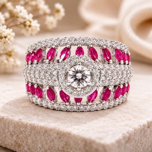 Può includere: Un ampio anello color argento con una pietra centrale rotonda taglio brillante circondata da un alone di piccole pietre chiare. La fascia è decorata con due file di pietre rosa vivace taglio marquise e diverse file di piccole pietre chiare.
