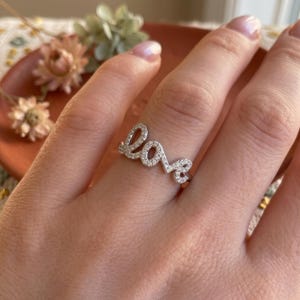 以下が含まれることがあります： 筆記体でloveという文字がデザインされたシルバー調の指輪。文字の表面全体に小さな透明な石が埋め込まれています。指に装着された状態で、精巧な金属細工と輝く石のセッティングがはっきりと確認できます。