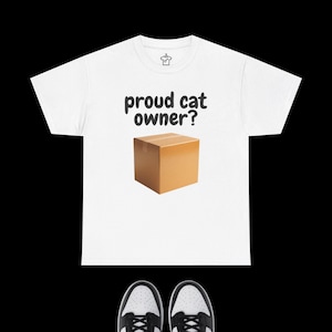 May include: A white short-sleeved t-shirt featuring the black text "proud cat owner?" printed above a graphic of a brown cardboard box. The shirt has a small hanger icon logo on the inside neck label area and is displayed above two black and white sneakers.
