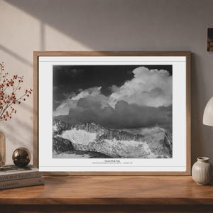 May include: A black and white landscape photograph in a wooden frame featuring a mountain range under large, dramatic clouds. The print includes the text "Clouds White Pass" and "East Peak Clouds, Yosemite Wilderness, California - Ansel Adams, 1948" at the bottom.