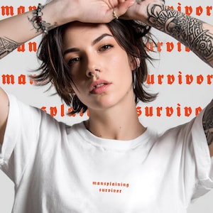 Könnte beinhalten: Ein weißes T-Shirt mit den Worten "mansplaining survivor" in roter gotischer Schrift. Der Hintergrund zeigt die Wörter "man" und "survivor" in einem sich wiederholenden Muster. Die Person im Bild hat Tätowierungen und trägt silberne Creolen.