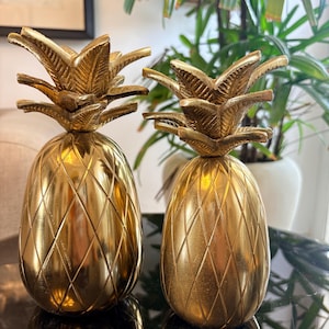 Op de afbeelding: Twee decoratieve ananasbeeldjes gemaakt van gepolijst goudkleurig metaal. Elk stuk heeft een gestructureerd lichaam met een ruitpatroon en een gedetailleerde, stekelige bladerkroon aan de bovenkant. De oppervlakken zijn glad en reflecterend.
