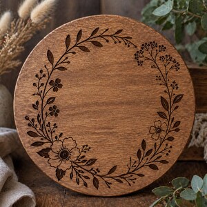 Könnte beinhalten: Ein runder Servierteller aus Holz mit einem floralen Kranzdesign. Der Teller ist aus hellbraunem Holz gefertigt und weist ein detailliertes, dunkelbraunes, graviertes Blumenmuster am Rand auf. Das Design umfasst Blumen, Blätter und Zweige.