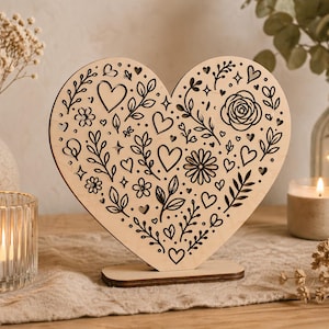 Könnte beinhalten: Holzdekoration in Herzform mit detaillierten schwarzen Blumen- und Herzmustern. Das Herz steht auf einem kleinen Sockel. Das Bild zeigt auch Kerzen, Vasen mit Trockenblumen und einen neutralen Hintergrund.