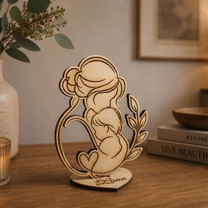 Puede incluir: Escultura de madera clara que representa a una madre y su hijo, con la palabra "Love" en una base en forma de corazón. La escultura presenta detalles recortados. Una vela en un vaso y un jarrón con vegetación están en el fondo.