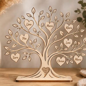 Könnte beinhalten: Ein hölzerner Familienstammbaum mit herzförmigen Blättern, auf denen jeweils ein Name steht. Der Baum ist aus hellem Holz gelasert und steht auf einem rechteckigen Sockel. Die Namen sind Lina, Mara, Klara, Leni, Chrissi, Marie, Pia und Sara.