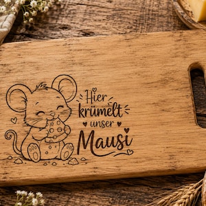 Puede incluir: Tabla de cortar de madera con asa, con un ratón de dibujos animados comiendo queso. El texto "Hier krümelt unser Mausi" está grabado en la tabla. La tabla es rectangular con un acabado de veta de madera natural.