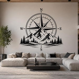 May include: Black and white wall art depicting a compass rose design with a mountain range and pine trees. The compass rose has the letters N, E, S, and W. The artwork is mounted on a neutral-coloured wall in a modern living room.