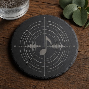 Puede incluir: Un posavasos redondo de pizarra negra con un diseño de nota musical. El diseño presenta una clave de sol y un patrón de onda de sonido dentro de círculos concéntricos y líneas punteadas. El posavasos está sobre una superficie de madera.