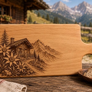 May include: Wooden cutting board with a handle, featuring an engraved mountain cabin scene with edelweiss flowers. The board is light brown and has a natural wood grain. A leather cord is attached to the handle. The board is approximately 15 inches long.