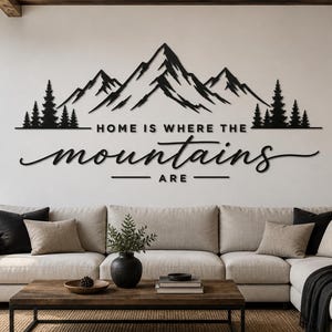 May include: Black metal wall art depicting a mountain range with trees and the text "HOME IS WHERE THE mountains ARE". The artwork is above a beige sofa in a living room. The room has a wooden coffee table.