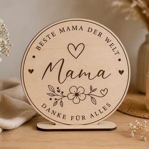 Könnte beinhalten: Runder Holzschild mit den Worten "Beste Mama der Welt" und "Danke für Alles" in dekorativer Schrift graviert. Das Schild zeigt ein Herz- und Blumendesign. Das Schild steht auf einem Holzständer.