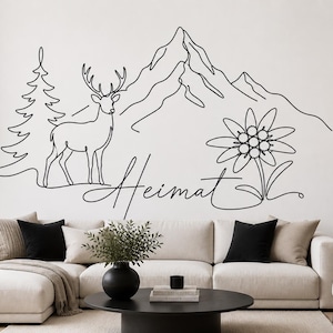 Könnte beinhalten: Schwarze Wanddekoration mit Linienkunst, die eine Bergkette, ein Reh, einen Baum und eine Blume darstellt. Das Wort "Heimat" ist in Schreibschrift geschrieben. Das Kunstwerk wird über einem beigen Sofa mit schwarzen und beigen Kissen gezeigt.