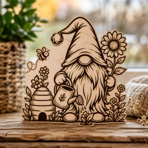 Könnte beinhalten: Gartenwichtel aus Holz mit langem Bart und Hut, der eine Gießkanne und eine Sonnenblume hält. Eine Biene und ein Bienenstock sind ebenfalls im Design enthalten. Der Wichtel ist von Blumen umgeben, alle aus Holz gelasert.
