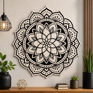 Könnte beinhalten: Ein großes, rundes Wandkunstwerk mit Mandala-Motiv aus Holz. Das Design zeigt ein symmetrisches Muster aus schwarzen und cremefarbenen floralen und geometrischen Formen. Das Kunstwerk ist an einer hellen Wand befestigt und erzeugt einen auffälligen Kontrast.