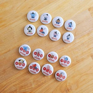 May include: A selection of white circular badges featuring cartoon characters and the word "ARIRANG" in red. The badges are displayed on a light brown wooden surface.