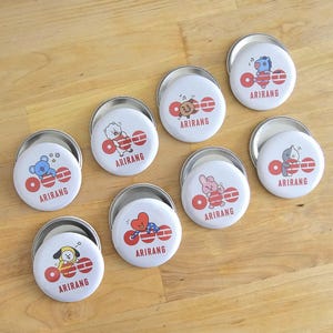 Puede incluir: Ocho chapas redondas con alfiler que muestran varios personajes de dibujos animados coloridos. Cada chapa tiene un fondo blanco, un diseño gráfico rojo y el texto ARIRANG impreso en rojo. Las chapas están dispuestas de forma dispersa.