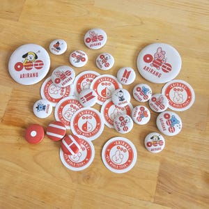 Op de afbeelding: Een verzameling witte en rode ronde knopen en stickers met cartoonfiguren en het woord "ARIRANG". Sommige knopen hebben de tekst "BTS WORLD TOUR". De items zijn verspreid over een houten oppervlak.