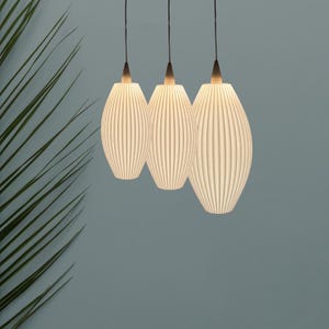 Könnte beinhalten: Drei Pendelleuchten mit geripptem, cremefarbenem, ovalem Design. Die Leuchten hängen an schwarzen Kabeln und strahlen ein warmes Licht aus. Der Hintergrund ist ein gedämpftes Blau-Grau mit einer grünen Pflanze in der Ecke.