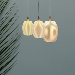 Könnte beinhalten: Drei Pendelleuchten mit geripptem, cremefarbenem, ovalem Design. Jede Leuchte hängt an einem schwarzen Kabel und hat einen Holzaufsatz. Die Leuchten sind beleuchtet und strahlen ein warmes Licht aus. Im Hintergrund ist ein grünes Blatt zu sehen.