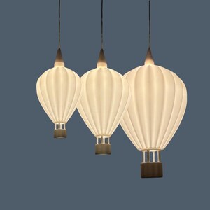 Könnte beinhalten: Drei Pendelleuchten in Form von Heißluftballons. Die weißen, gerippten Schirme hängen an dunklen Schnüren. Der Sockel jedes Ballons ist dunkler, der Hintergrund ist ein gedämpftes Blau.