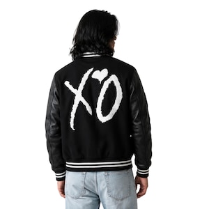 May include: A black varsity jacket featuring long black sleeves and a white graphic on the back that displays the letters XO with a small heart symbol above the O. The jacket has a ribbed collar, cuffs, and hem with white stripes.