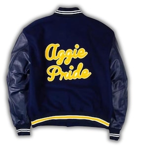 Pode incluir: Uma jaqueta universit&aacute;ria azul-marinho com mangas compridas de couro e gola, punhos e bainha canelados com listras brancas e amarelas. As costas exibem o texto Aggie Pride em letras cursivas grandes e amarelas com contorno branco.
