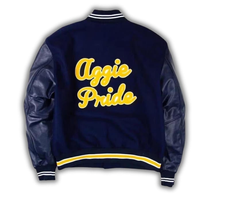 Pode incluir: Uma jaqueta universit&aacute;ria azul-marinho com mangas compridas de couro e gola, punhos e bainha canelados com listras brancas e amarelas. As costas exibem o texto Aggie Pride em letras cursivas grandes e amarelas com contorno branco.
