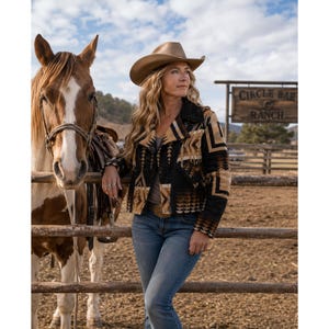 May include: A tan cowboy hat and a black, tan, and brown patterned jacket with a geometric design, worn with blue jeans. The jacket features a wide collar and long sleeves, standing next to a brown and white horse by a wooden fence sign.