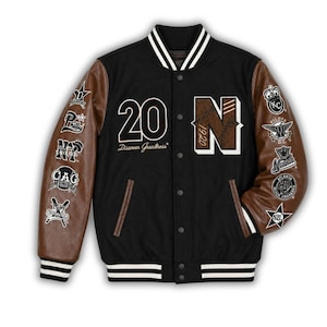 Peut inclure: Veste de style universitaire noire avec des manches en cuir marron. Elle présente le chiffre "20" et la lettre "N" avec "1920" sur le devant. Des écussons ornent les manches. La veste a des bordures blanches et noires rayées au col, aux poignets et à l'ourlet. Le texte "Discover Greatness" est également visible.