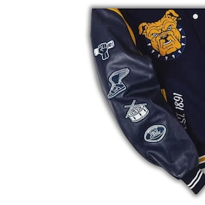 Pode incluir: Um casaco varsity azul-marinho com punhos canelados &agrave;s riscas amarelas e brancas. A manga apresenta emblemas bordados a branco de um punho, um t&eacute;nis alado, um tambor e uma bola de futebol americano. O peito exibe uma cabe&ccedil;a de bulldog amarela e o texto EST. 1891.