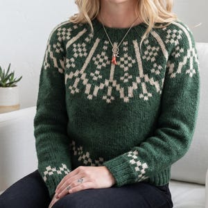 May include: A green knitted jumper featuring a cream geometric design across the chest and sleeves. The jumper has a round neck and long sleeves. A silver necklace with a red pendant is visible.