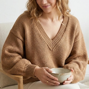 May include: A tan, oversized V-neck sweater with ribbed cuffs and neckline. The sweater is made of a thick knit material. A person is holding a small, light-colored ceramic bowl.