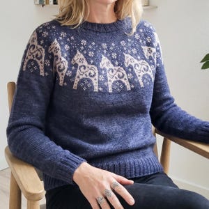 May include: A navy blue knitted sweater with a crew neck and long sleeves. The sweater has a pattern of light beige horses and snowflake designs across the chest and shoulders. The ribbed cuffs and hem provide a snug fit. The sweater is made of a soft, knitted material.