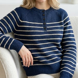 May include: Navy blue knit sweater with horizontal cream stripes. The sweater has a crew neck, long sleeves, and a button-up placket. The model is wearing the sweater while sitting on a light-colored chair.