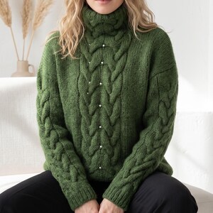 Könnte beinhalten: Ein grüner Zopfmuster-Rollkragenpullover mit Perlenakzenten. Der Pullover hat ein dickes, strukturiertes Strickdesign mit hohem, umgeschlagenem Kragen. Das Zopfmuster verläuft über die Vorderseite und die Ärmel. Kleine, weiße Perlen sind in der Mitte des Pullovers aufgenäht.