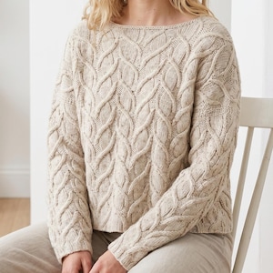 May include: Cream-colored cable-knit sweater with a boat neck. The sweater features a detailed, textured pattern across the front, sleeves, and shoulders. The knitwear is a neutral color, suitable for various outfits and occasions. The sleeves are long and the fit is relaxed.