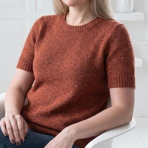 Peut inclure: Un pull en tricot rouille à manches courtes. Le pull a un col rond et un aspect texturé. Les manches s'arrêtent juste au-dessus du coude. La personne porte un jean bleu et est assise sur une chaise blanche.