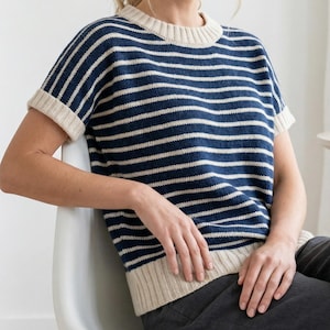 Peut inclure: Un pull à manches courtes rayé bleu marine et crème. Le pull présente des rayures horizontales, un col rond et des revers aux manches. Le bas et l'encolure sont de couleur crème. La personne est assise sur une chaise blanche.