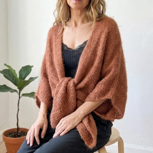 May include: A terracotta-coloured knitted shawl with a draped design. The shawl is tied at the front, creating a layered effect. The soft, textured knitwear is a warm, cosy accessory. The shawl is worn over a black top and black trousers.