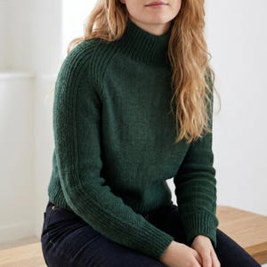 May include: A forest green turtleneck sweater with ribbed detailing on the shoulders and sleeves. The sweater has a textured knit pattern and a fitted silhouette. The model is wearing dark blue jeans.