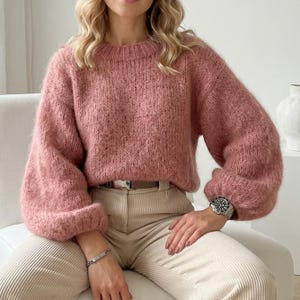 Könnte beinhalten: Ein staubrosa farbener Pullover mit rundem Ausschnitt und langen, voluminösen Ärmeln. Der Pullover ist aus einem strukturierten Strickstoff gefertigt. Das Model trägt eine silberne Uhr und ein Armband. Die Hose ist Cord.