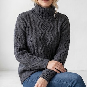 May include: A dark gray cable-knit turtleneck sweater with intricate patterns. The sweater features a high, ribbed collar and is paired with blue jeans. The knitwear has a textured appearance, with detailed cable and diamond designs.